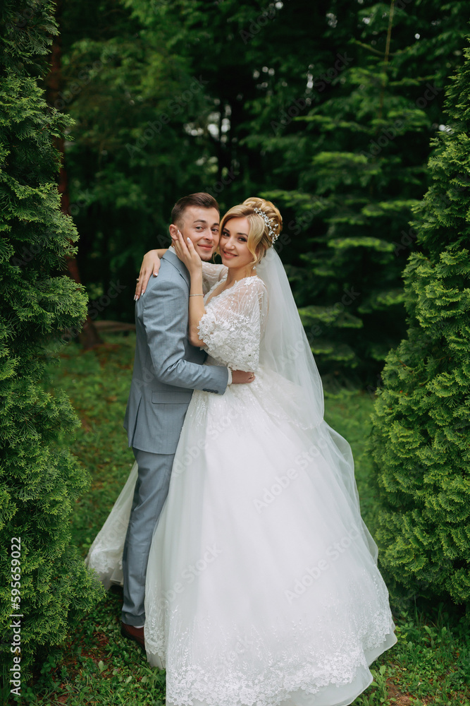 Portrait of the bride and groom standing on the background of green trees, embracing. The bride smiles sincerely. Stylish groom. Fashion and style. Beautiful bride Portrait of the bride and groom standing on the background of green trees, embracing. The bride smiles sincerely. Stylish groom. Fashion and style. Beautiful bride