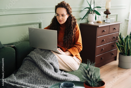 Relaxed young woman sitting on sofa holding mobile phone using cellphone and laptop technology doing online ecommerce shopping, texting messages relaxing on couch in cozy living room home interior.