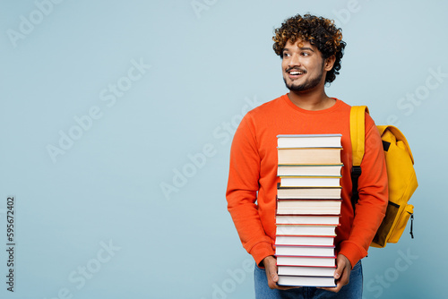 Young teen Indian boy student wear casual clothes backpack bag hold in hand pile of many books look aside isolated on plain pastel light blue cyan background. High school university college concept.