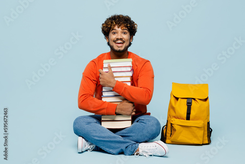Full body surprised excited young teen Indian boy student wear casual clothes sit near backpack bag hold pile stack of many books isolated on plain pastel blue background. High school college concept. © ViDi Studio