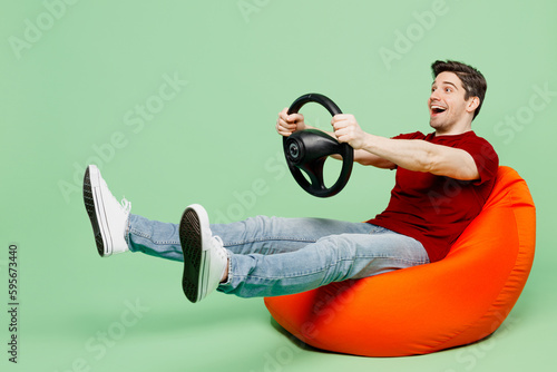 Full body surprised happy young man wear red t-shirt casual clothes sit in bag chair hold steering wheel driving car isolated on plain pastel light green background studio portrait. Lifestyle concept photo