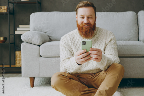 Young happy ginger man wear casual clothes hold in hand use mobile cell phone sits near grey sofa couch stay at home hotel flat rest relax spend free spare time in living room indoor. Lounge concept.
