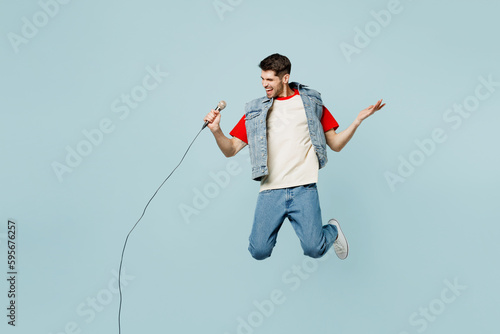 Full body singer young man he wear denim vest red t-shirt casual clothes jump high sing song in microphone at karaoke club isolated on plain pastel light blue cyan background studio Lifestyle concept