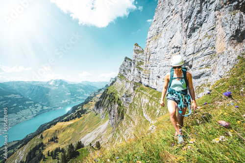 Sportive woman hiking on flowery trail next to steep rock wall with scenic view on Walensee and Churfürsten mountain range in the background. Schnürliweg, Walensee, St. Gallen, Switzerland, Europe.