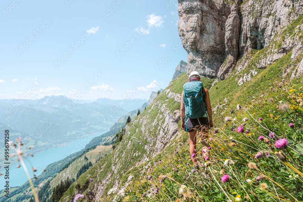 Back View Of Young Woman Hiking On Flowery Trail Next To Steep Rock Wall With Scenic View On Back View Of Young Woman Hiking On Flowery Trail Next To Steep Rock Wall With Scenic View On