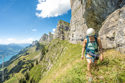 Front view of sporty woman hiking on meadow below steep rock wall with scenic view on lake Walensee and the swiss alps. Schnürliweg, Walensee, St. Gallen, Switzerland, Europe.