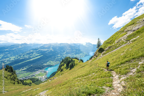 Athletic woman hikes on flowery trail on steep meadow with panoramic view on lake Walensee and the swiss alps. Schnürliweg, Walensee, St. Gallen, Switzerland, Europe.