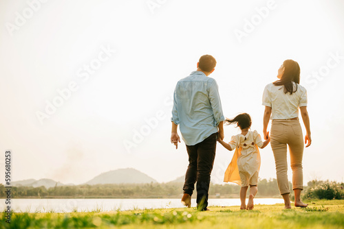 A young Asian girl holding hands with her parents and walking in a green field, with a sunny sky and a feeling of joy and happiness, Happy Family day, back view