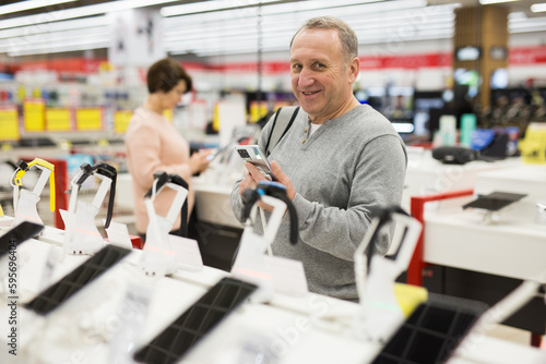 Mature caucasian man looking at camera and smiling while choosing smartphone in electronic shop.