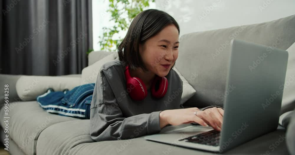 Excited asian woman typing on laptop while laying on sofa, communicating with friends Excited asian woman typing on laptop while laying on sofa, communicating with friends