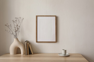 Empty wooden picture frame mockup hanging on beige wall background. Boho shaped vase, dry flowers on table. Cup of coffee, old books. Working space, home office. Art, poster display. Modern interior.