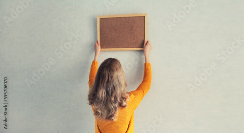 Fotografie Young woman decorating interior, hanging blank a photo frame mockup on white wal