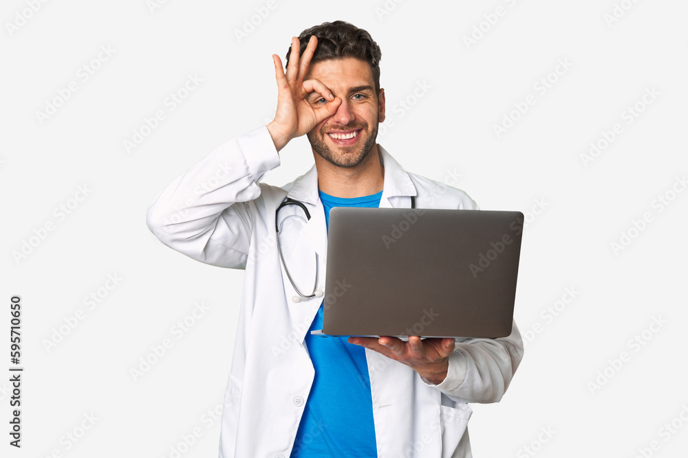 Young doctor man holding a laptop excited keeping ok gesture on eye. Young doctor man holding a laptop excited keeping ok gesture on eye.