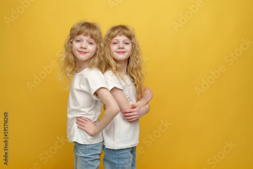 A portrait of twin girls. Two beautiful and positive curly blonde sisters who are 6 or 7 years old, wearing white t-shirts and casual jeans, standing back to back on a yellow background.