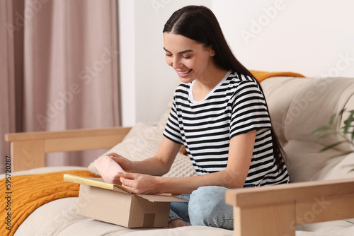 Happy young woman opening parcel on sofa at home. Internet shopping