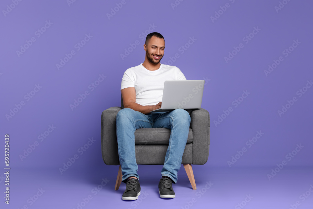 Smiling young man working with laptop in armchair on lilac background Smiling young man working with laptop in armchair on lilac background