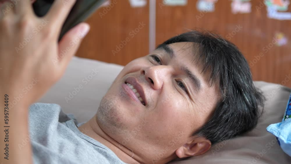 Asian adult man holding and looking at smartphone or mobile with happy smiling face on bed in bedroom. Concept of technology, communication, connection. Asian adult man holding and looking at smartphone or mobile with happy smiling face on bed in bedroom. Concept of technology, communication, connection.