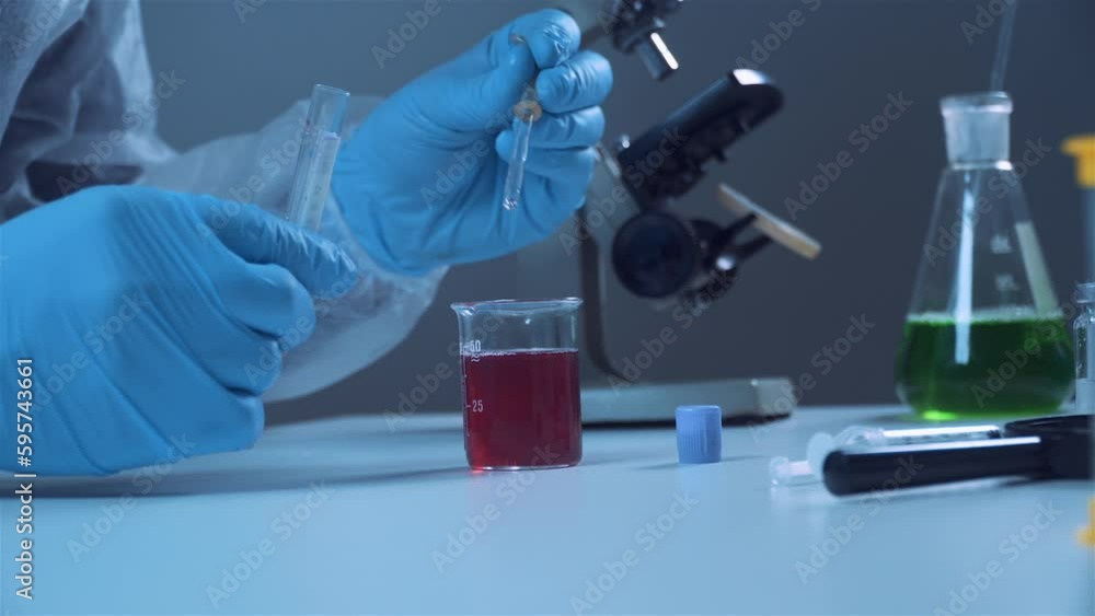 Close-up of hand of a chemical laboratory worker in protective white clothing and blue latex gloves takes a sample of a red liquid with a pipette. Scientist with pipette analysis red colored liquid Close-up of hand of a chemical laboratory worker in protective white clothing and blue latex gloves takes a sample of a red liquid with a pipette. Scientist with pipette analysis red colored liquid