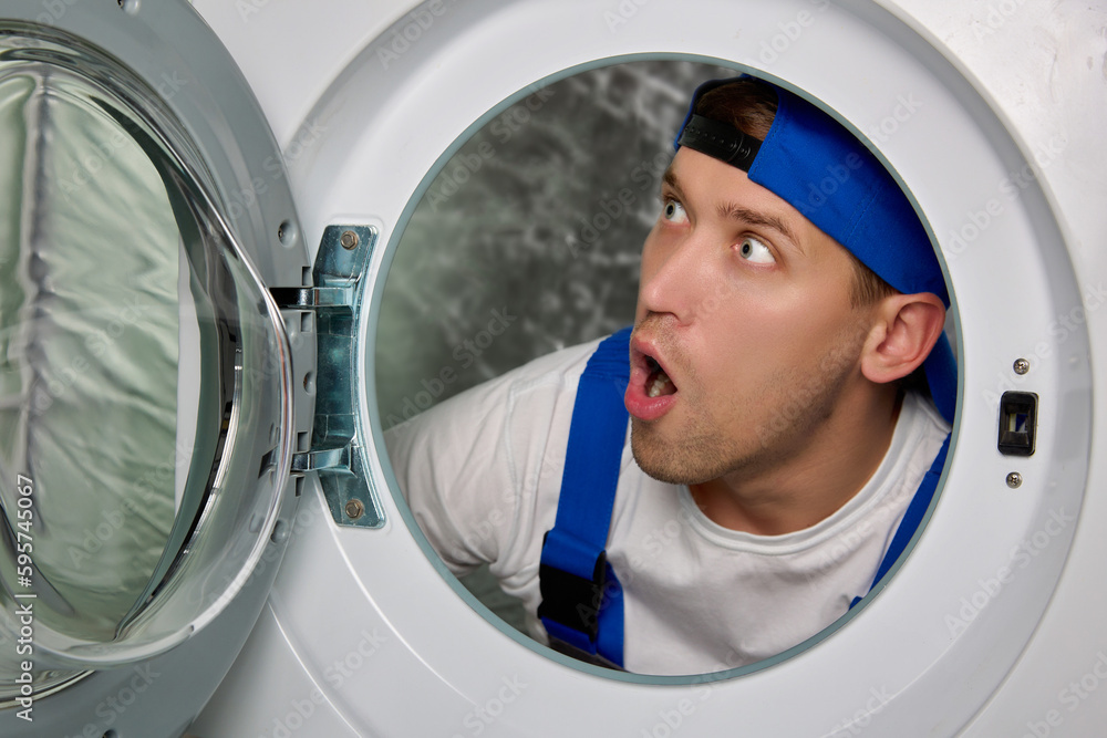 With a surprised look and with his mouth open, a young man in a uniform from the drum of a washing machine looks. Experienced working plumber shocked by the scale of the breakdown and says wow With a surprised look and with his mouth open, a young man in a uniform from the drum of a washing machine looks. Experienced working plumber shocked by the scale of the breakdown and says wow