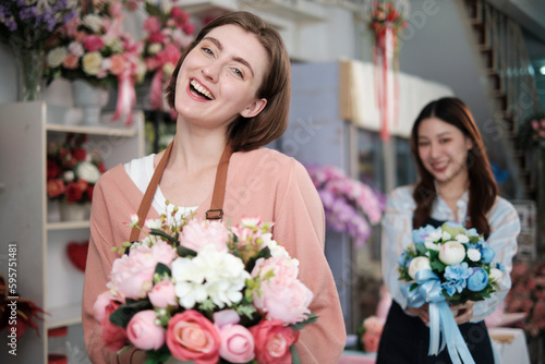 Young beautiful White female florist worker with bunch of blossoms and her colleague smile and look at camera at colorful flower shop in Valentine, small business occupation, happy SME entrepreneur.