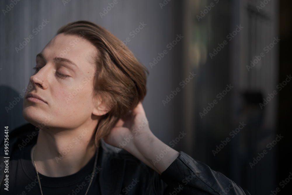 A genderfluid model (he, she, they) wears a black leather jacket and stands in front of a gray wall, running their hand through their hair. A genderfluid model (he, she, they) wears a black leather jacket and stands in front of a gray wall, running their hand through their hair.