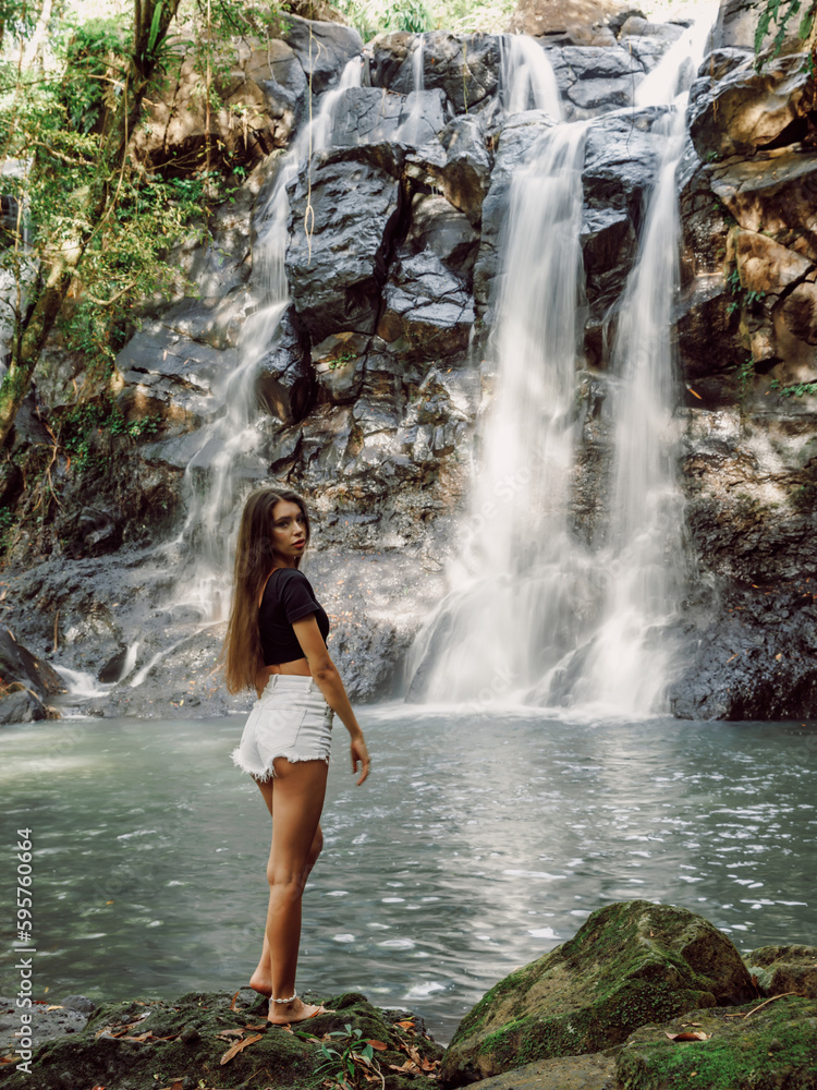Beautiful slim woman on waterfall in Bali. Traveler girl posing on river with waterfall
