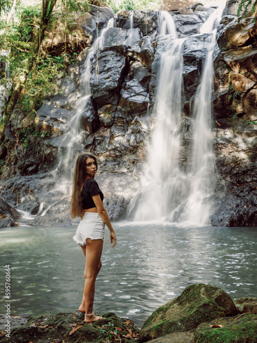 Beautiful slim woman on waterfall in Bali. Traveler girl posing on river with waterfall