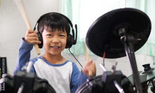 Asian child boy hold drumstick to play electronic drum with headphone and happy smiling face at home. Concept of music learning, happy relaxing time, freedom, little kid musician.