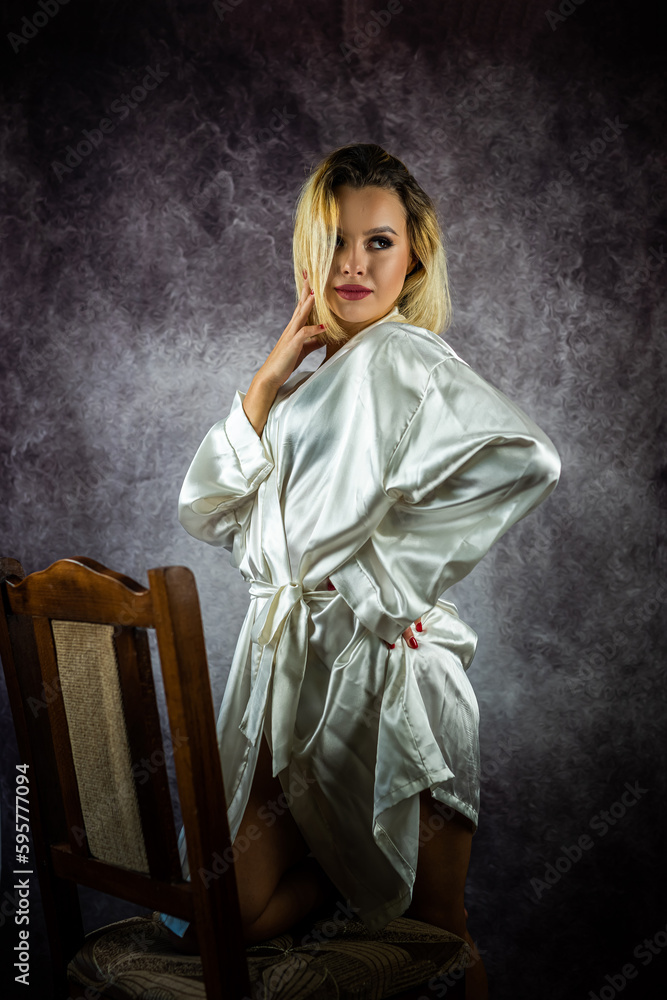 Beautiful adult sexy woman sitting on a chair in a white silk robe posing in the studio. Beautiful adult sexy woman sitting on a chair in a white silk robe posing in the studio.
