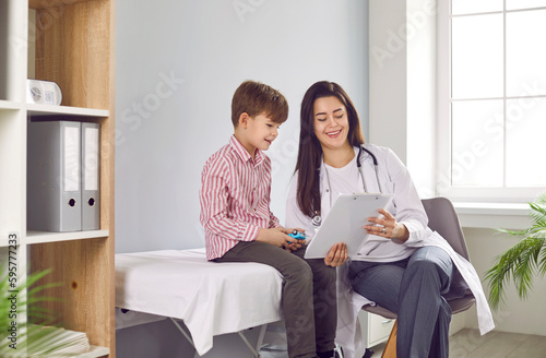 Portrait of a friendly smiling female child doctor pediatrician showing report file with appointment to a little boy patient sitting on the couch during medical examination in clinic. photo