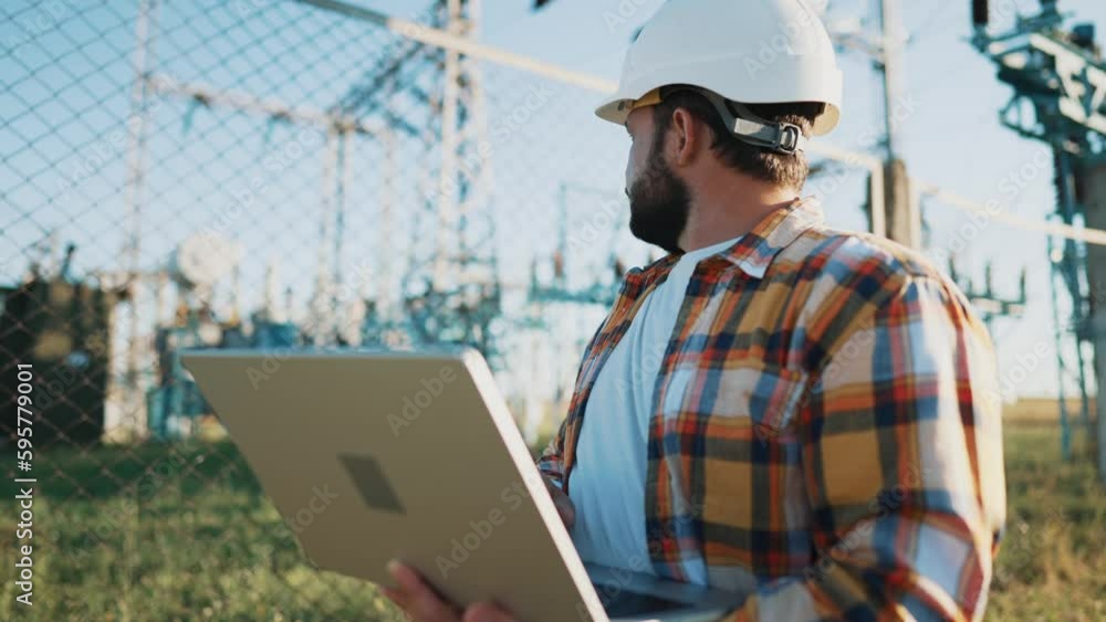 Male engineer using laptop computer to online work in wheat field thinks and rubs his beard. Man in white helmet stares intently at screen and typing on keyboard against high telecommunications tower. Male engineer using laptop computer to online work in wheat field thinks and rubs his beard. Man in white helmet stares intently at screen and typing on keyboard against high telecommunications tower.