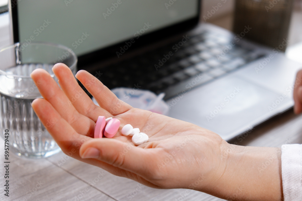 Woman taking Daily vitamins white working on laptop Organizer weekly shots Closeup of medical pill box with doses of tablets for daily take medicine with white pink drugs and capsules. Workplace Woman taking Daily vitamins white working on laptop Organizer weekly shots Closeup of medical pill box with doses of tablets for daily take medicine with white pink drugs and capsules. Workplace
