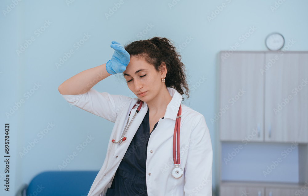 Exhausted female doctor in medical clothes touches temple eyes closed