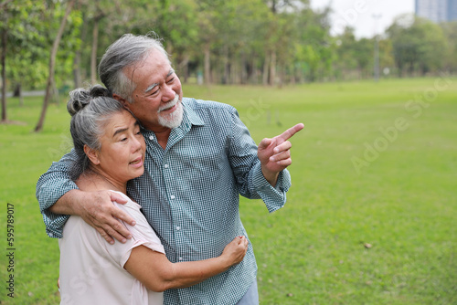 Happy asian senior man and woman walking and hugging while pointing something with picnic basket in garden outdoor. Lover couple going to picnic at the park. Happiness marriage lifestyle concept.