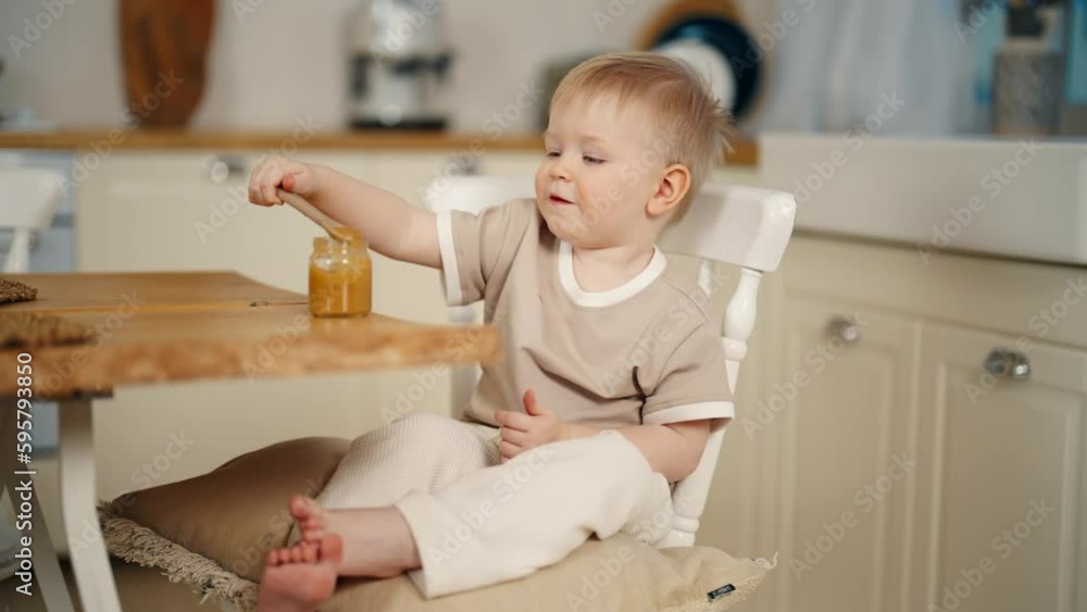 Smiling baby girl eats puree holds spoon in hand by herself sitting on chair at table in kitchen at home. Adorable little kid takes fruit or vegetable puree from jar. Babyhood, childhood concept. Smiling baby girl eats puree holds spoon in hand by herself sitting on chair at table in kitchen at home. Adorable little kid takes fruit or vegetable puree from jar. Babyhood, childhood concept.