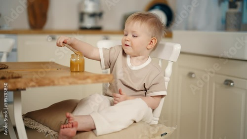 Smiling baby girl eats puree holds spoon in hand by herself sitting on chair at table in kitchen at home. Adorable little kid takes fruit or vegetable puree from jar. Babyhood, childhood concept. photo