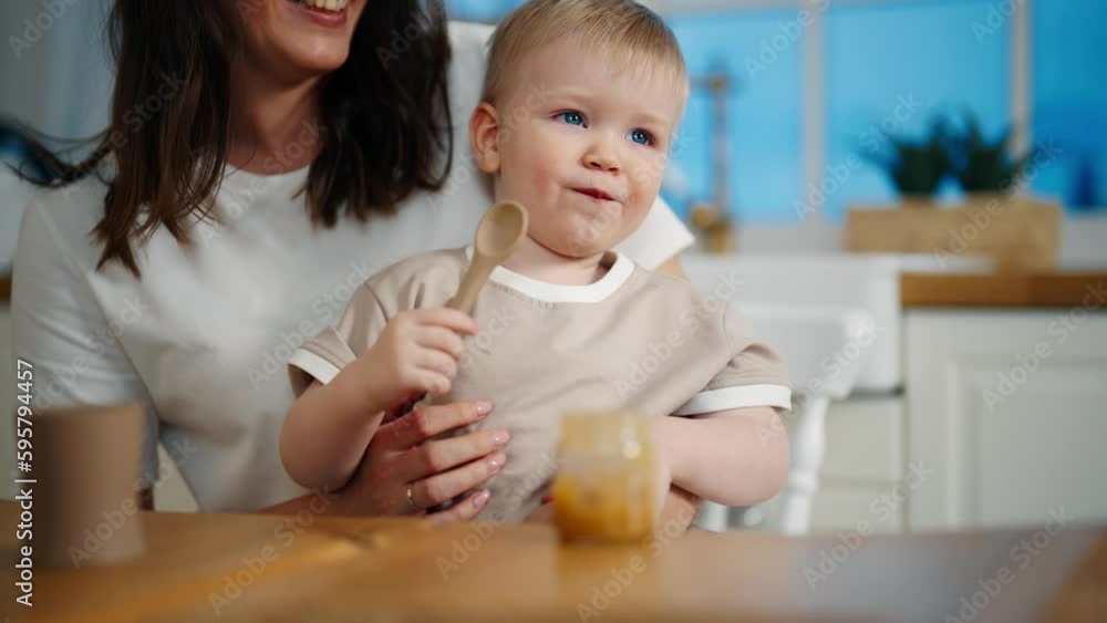 Happy smiling positive mom looks on baby girl eats herself fruit or vegetable puree from jar with spoon sits on kitchen at home. Babyhood, childhood, maternity leave, rising child motherhood concept. Happy smiling positive mom looks on baby girl eats herself fruit or vegetable puree from jar with spoon sits on kitchen at home. Babyhood, childhood, maternity leave, rising child motherhood concept.