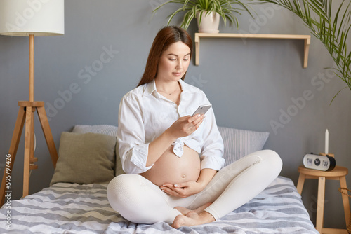 Brown haired beautiful cute future mother using mobile phone, pregnant woman with bare belly sitting in bed browsing internet, planning maternity home.