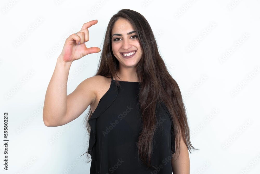 young brunette woman wearing black blouse over white studio background smiling and confident gesturing with hand doing small size sign with fingers looking and the camera. Measure concept young brunette woman wearing black blouse over white studio background smiling and confident gesturing with hand doing small size sign with fingers looking and the camera. Measure concept