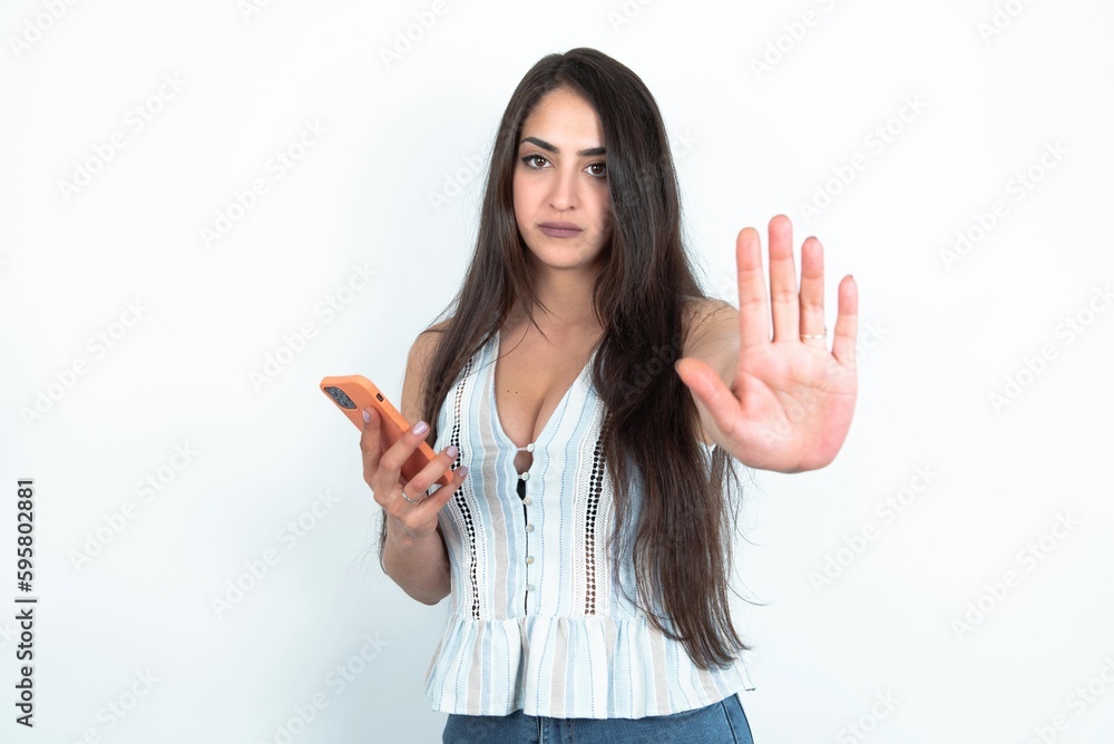 young brunette woman wearing white tank top over white studio background using and texting with smartphone with open hand doing stop sign with serious and confident expression, defense gesture young brunette woman wearing white tank top over white studio background using and texting with smartphone with open hand doing stop sign with serious and confident expression, defense gesture