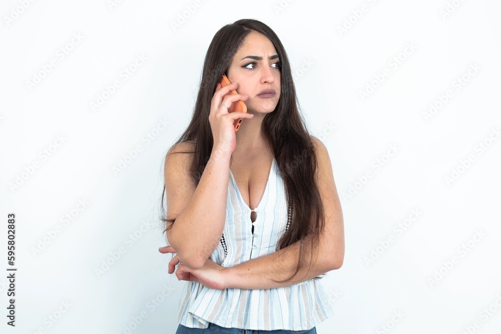 Sad young brunette woman wearing white tank top over white studio background talking on smartphone. Communication concept. Sad young brunette woman wearing white tank top over white studio background talking on smartphone. Communication concept.