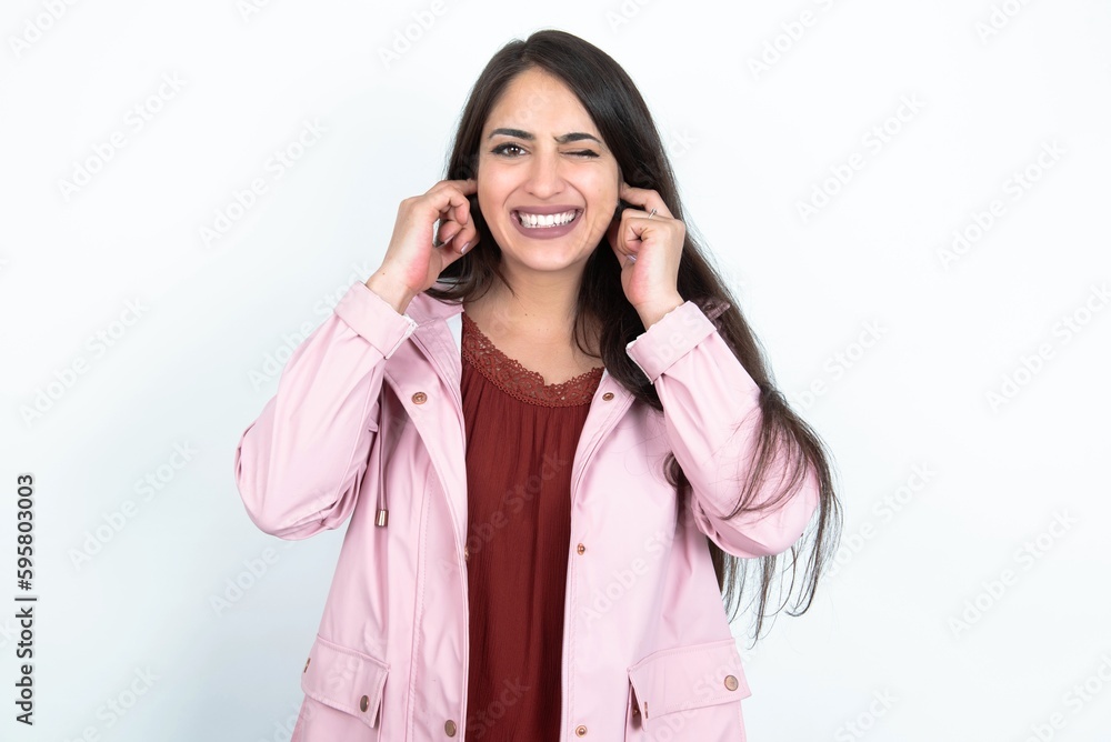 Happy young brunette woman wearing pink raincoat over white studio background ignores loud music and plugs ears with fingers asks to turn off sound Happy young brunette woman wearing pink raincoat over white studio background ignores loud music and plugs ears with fingers asks to turn off sound
