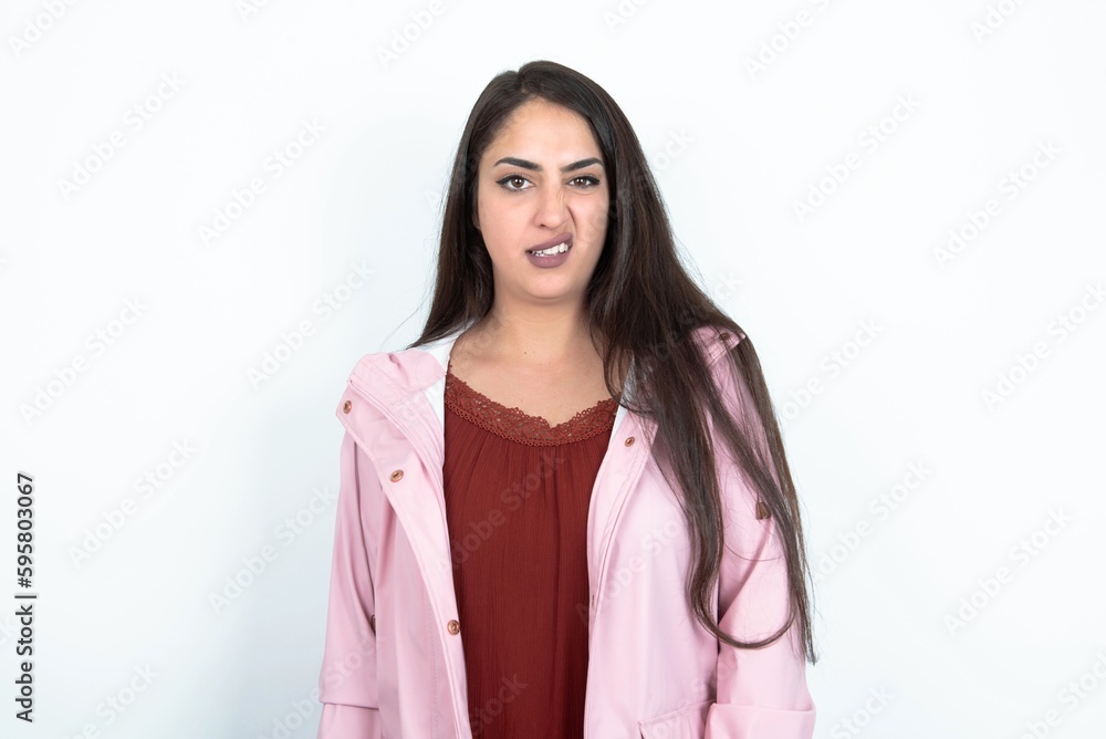 Portrait of dissatisfied young brunette woman wearing pink raincoat over white studio background smirks face, purses lips and looks with annoyance at camera, discontent hearing something unpleasant Portrait of dissatisfied young brunette woman wearing pink raincoat over white studio background smirks face, purses lips and looks with annoyance at camera, discontent hearing something unpleasant