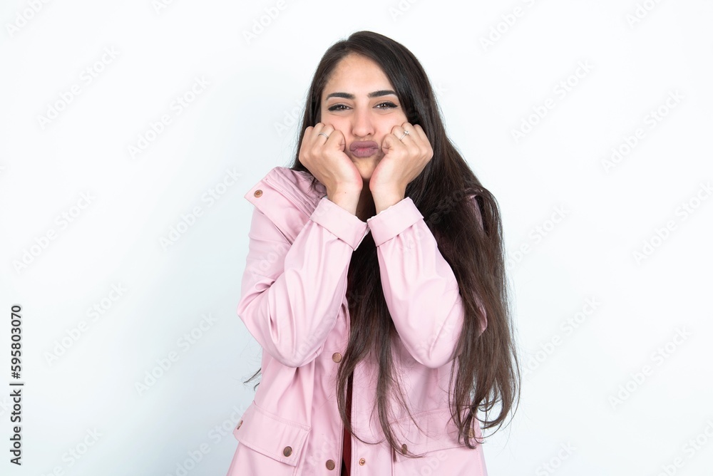 young brunette woman wearing pink raincoat over white studio background with surprised expression keeps hands under chin keeps lips folded makes funny grimace young brunette woman wearing pink raincoat over white studio background with surprised expression keeps hands under chin keeps lips folded makes funny grimace