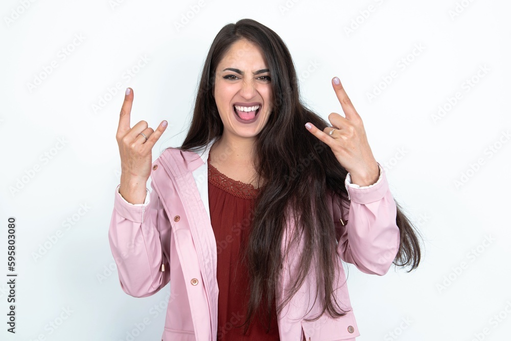 young brunette woman wearing pink raincoat over white studio background makes rock n roll sign looks self confident and cheerful enjoys cool music at party. Body language concept.