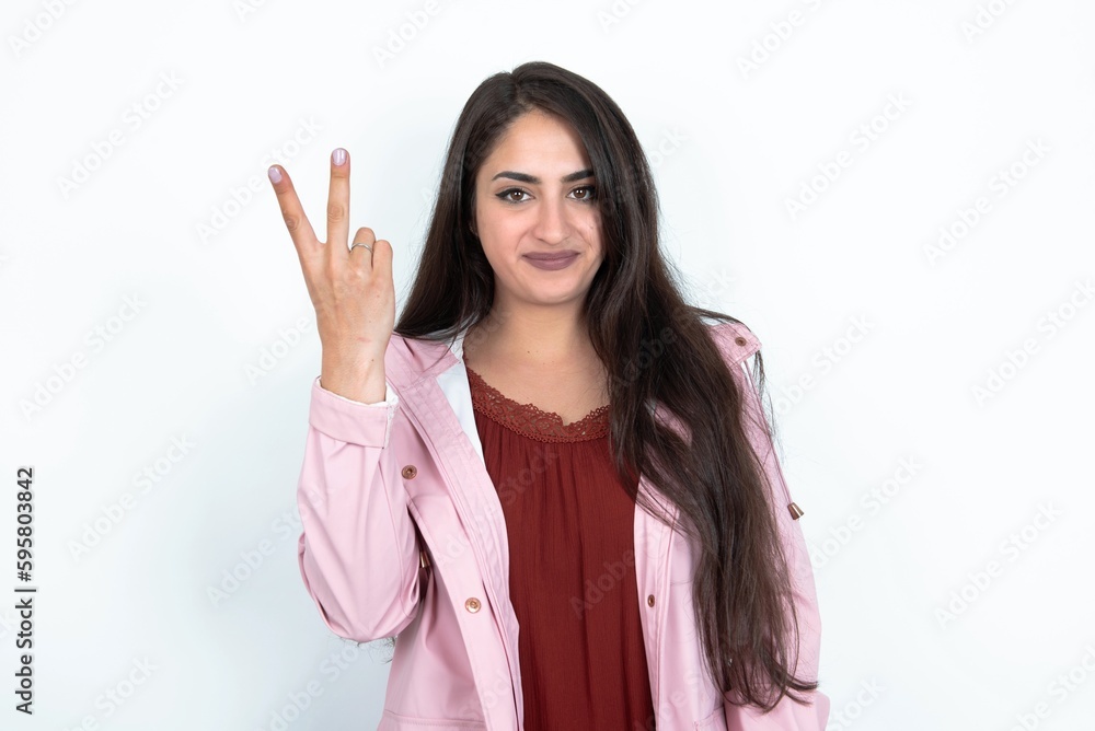 young brunette woman wearing pink raincoat over white studio background smiling and looking friendly, showing number two or second with hand forward, counting down young brunette woman wearing pink raincoat over white studio background smiling and looking friendly, showing number two or second with hand forward, counting down