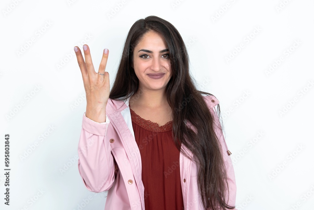 young brunette woman wearing pink raincoat over white studio background smiling and looking friendly, showing number three or third with hand forward, counting down young brunette woman wearing pink raincoat over white studio background smiling and looking friendly, showing number three or third with hand forward, counting down