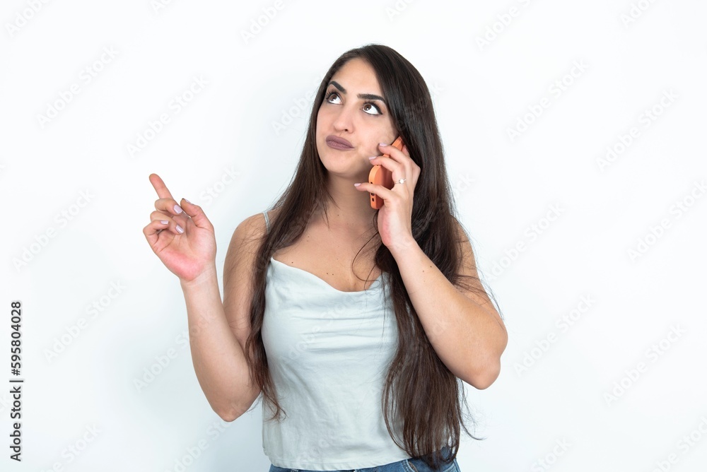 young brunette woman wearing white tank top over white studio background speaks on mobile phone spends free time indoors calls to friend. young brunette woman wearing white tank top over white studio background speaks on mobile phone spends free time indoors calls to friend.