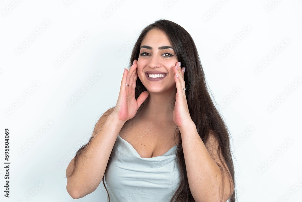 Happy young brunette woman wearing white tank top over white studio background touches both cheeks gently, has tender smile, shows white teeth, gazes positively straightly at camera,