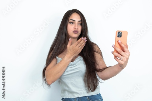 young brunette woman wearing white tank top over white studio background blows air kiss at camera of smartphone and takes selfie, sends mwah via online call.